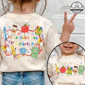 May include: A cream-colored t-shirt featuring a colorful graphic print of school supplies with faces, including a pencil, apple, globe, and notebook. The text reads It's a Good day for Learning in multi-colored letters surrounded by small stars.