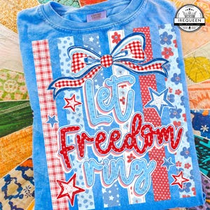Puede incluir: Camiseta azul claro con un diseño patriótico. La camiseta presenta el texto "Let Freedom Ring" en rojo y azul, junto con estrellas y un lazo. El diseño incorpora cuadros rojos y blancos y lunares azules.