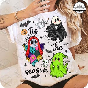 May include: A white t-shirt featuring a graphic print of three ghosts, a pumpkin, a skull, and bats. The text reads 'tis the season'. The ghosts are depicted in black, red, and green, with spider web accents surrounding the illustrated characters.
