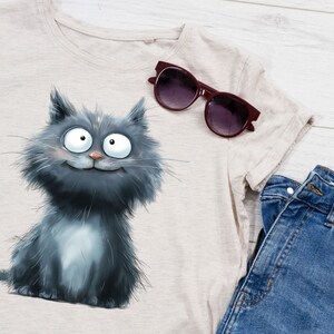 May include: A light beige t-shirt featuring a cartoon cat illustration in shades of gray. The cat has large, expressive eyes and a smiling face. A pair of burgundy sunglasses and blue denim jeans are also visible.