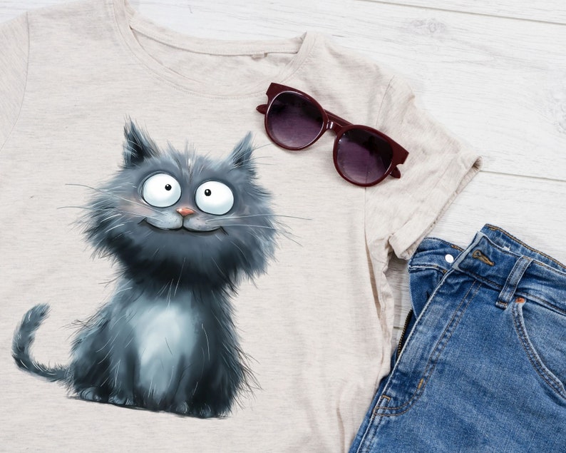 May include: A light beige t-shirt featuring a cartoon cat illustration in shades of gray. The cat has large, expressive eyes and a smiling face. A pair of burgundy sunglasses and blue denim jeans are also visible.