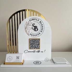May include: A white acrylic display stand with gold detailing. The stand displays the text "Simply Blessed Designs" and a QR code. A business card holder and a wireless charging pad are also present. The phrase "Connect with us!" is written below the QR code.