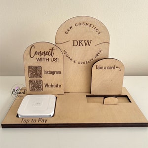 May include: A wooden point-of-sale display with multiple sections. It features the text "DKW Cosmetics, Vegan & Cruelty Free", "Connect With Us!" with Instagram and website QR codes, and "Take a card". A white card reader is in the base.