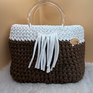 May include: A handmade crochet tote bag with a clear acrylic handle. The bag has a brown base and a white top, with white fringe and a small wooden tag. The bag is on a white fluffy surface.