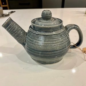 May include: A handcrafted, grey ceramic teapot with a lid and handle. The teapot has a spout and a round knob on top of the lid. The exterior features horizontal, textured lines. A small tag is attached to the handle with twine. The teapot is on a white surface.