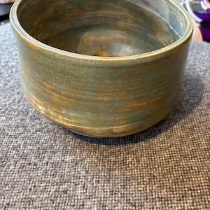 May include: A handmade, circular ceramic bowl with a broad opening. The bowl has a textured surface with a mix of green, brown, and blue tones. The pottery item is suitable for serving or display.