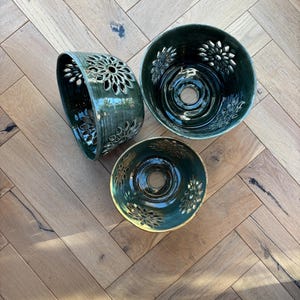 May include: Three dark green ceramic bowls with floral cut-out patterns. The bowls have a glossy finish and a central circular opening. They are displayed on a light brown wooden surface.