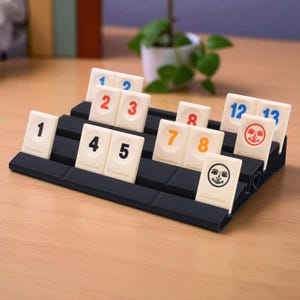 Könnte beinhalten: Ein schwarzer Rummikub-Kartenhalter mit weißen Kacheln, die Zahlen und Gesichter zeigen. Die Zahlen sind in Blau, Orange und Rot gehalten. Die Spielsteine sind in dem Halter auf einer hellbraunen Oberfläche angeordnet.