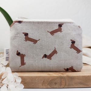 Könnte beinhalten: Eine kleine, rechteckige, beige Reißverschlusstasche mit einem sich wiederholenden Muster aus braunen Dackel-Hunden. Die Tasche ist aus einem strukturierten Stoff gefertigt und hat ein weißes Etikett mit dem Text "ELLEN'S BIRD". Die Tasche liegt auf einem Holzbrett.