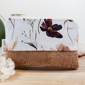 Könnte beinhalten: Eine kleine, rechteckige Geldbörse mit weißem Blumenmuster und Korkbasis. Die Geldbörse hat einen Reißverschluss und ein Etikett mit der Aufschrift "Ellen Spirit". Das florale Design umfasst burgunderfarbene Blumen und zarte Stiele.