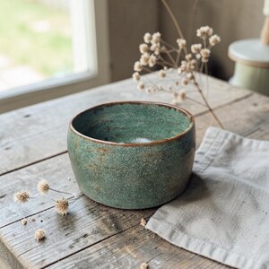 Peut inclure: Un bol rond en céramique vert-turquoise, avec une texture et un bord marron. Il est posé sur une table en bois vieilli, avec une serviette en lin et des fleurs séchées. L'ensemble évoque la décoration intérieure et les articles de cuisine artisanaux.