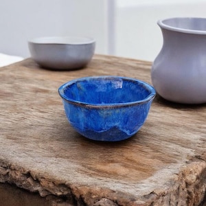 Peut inclure: Un petit bol en céramique fait à la main avec un glaçage bleu vibrant et une surface texturée. Le bol est posé sur une surface en bois rustique, avec deux autres pièces en céramique en arrière-plan. Le bol mesure environ 10 cm de diamètre.