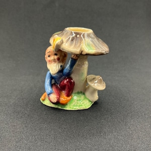 May include: A ceramic figurine of a gnome sitting next to a mushroom. The gnome wears a blue jacket, red pants, and has a white beard. The mushroom has a brown cap and a beige stem, with a smaller mushroom nearby.