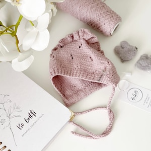 Può includere: Un cappellino da neonato in maglia rosa chiaro con bordo a balze e delicati dettagli traforati. Il cappellino ha lacci attaccati e una piccola etichetta. Sono inoltre mostrate due forcine per capelli in feltro grigio, una spoletta di filo e un taccuino con illustrazioni floreali.