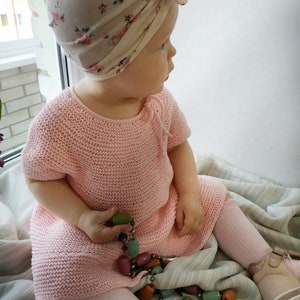 Puede incluir: Una ni&ntilde;a peque&ntilde;a con un vestido de punto rosa y un turbante con estampado floral. La ni&ntilde;a est&aacute; sentada en una manta a rayas y sostiene un collar de cuentas de colores.