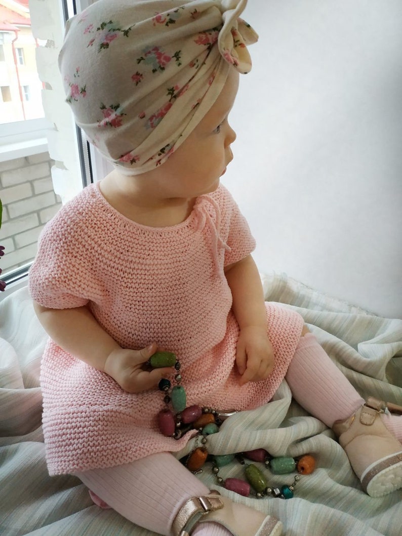 Puede incluir: Una ni&ntilde;a peque&ntilde;a con un vestido de punto rosa y un turbante con estampado floral. La ni&ntilde;a est&aacute; sentada en una manta a rayas y sostiene un collar de cuentas de colores.