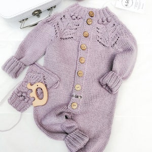 Op de afbeelding: Een lavendelkleurige gebreide baby romper met knoopsluiting aan de voorkant. De romper heeft een delicaat kantpatroon op de schouders en een klein houten speelgoedje bevestigd aan de zak.