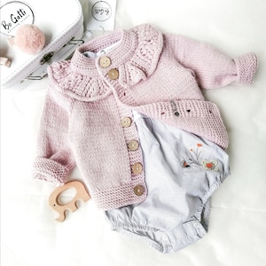 May include: A pink knitted cardigan with a ruffled collar and button closure. The cardigan is laid out on a white surface with a white and blue striped onesie and a wooden elephant toy.