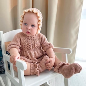 Puede incluir: Un bebé con un vestido de punto rosa y un gorro y calcetines a juego. El bebé está sentado en una mecedora blanca.