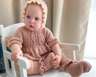 Op de afbeelding: Een baby die een roze gebreid jurkje draagt met bijpassende muts en sokken. De baby zit in een witte schommelstoel.