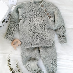 May include: A gray knitted baby sweater and pants set with a delicate lace pattern. The sweater has a button closure on the shoulder.