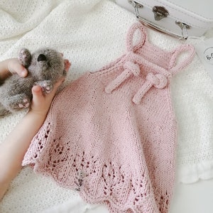 May include: A pink knitted baby dress with a bow at the neckline. The dress has a delicate lace trim around the bottom.