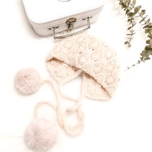 May include: A pale pink knitted baby hat with two fluffy pom-poms attached to the ties. The hat is laying on a white surface with a small white suitcase in the background.