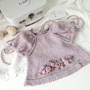 以下が含まれることがあります： 薄い紫色のニットのベビーワンピース。ピンクとブルーの刺繍の花が施されています。ワンピースは後ろにリボンが付いており、「Bo Gatti」というラベルが付いています。