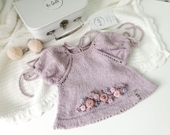 May include: A pale purple knitted baby dress with embroidered flowers in shades of pink and blue. The dress has a tie at the back and a label that says "Bo Gatti".