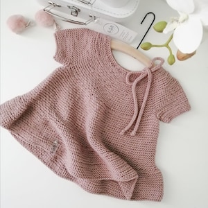 Puede incluir: Un vestido de beb&eacute; de punto rosa p&aacute;lido con mangas cortas y un lazo en la espalda. El vestido tiene una peque&ntilde;a etiqueta con el texto "La Gatti" en la parte delantera.