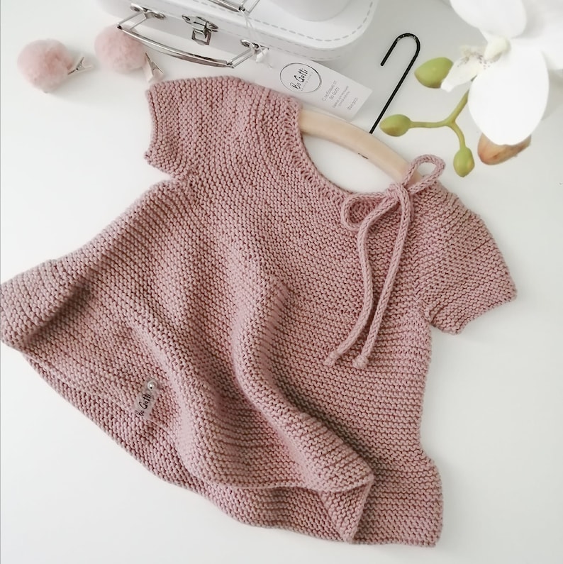 Puede incluir: Un vestido de beb&eacute; de punto rosa p&aacute;lido con mangas cortas y un lazo en la espalda. El vestido tiene una peque&ntilde;a etiqueta con el texto "La Gatti" en la parte delantera.