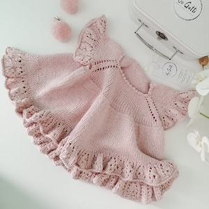 Pode incluir: Um casaquinho de bebê de malha rosa claro com bordas franzidas e detalhes delicados vazados. Dois grampos de cabelo de pompom rosa combinando e uma mala branca com o texto "Bo Gatti Knitting Studio" também são visíveis.