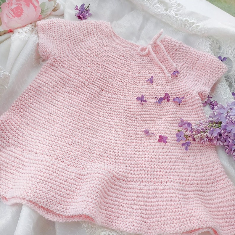 Puede incluir: Un vestido de beb&eacute; de punto rosa con mangas cortas y un lazo en el cuello. El vestido est&aacute; sobre un fondo de tela blanca con flores moradas.