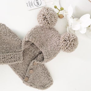 May include: A beige knitted baby hat with two fluffy pom-poms and a matching hooded cardigan with button closures.