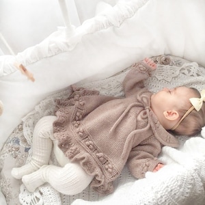 May include: A baby wearing a light brown knitted sweater with a ruffled hem and a bow on the back of the head. The baby is lying in a white wicker basket.
