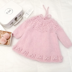 May include: A pink knitted baby dress with a lace pattern and pom-poms around the neckline and hem. The dress has a tie at the back of the neck.