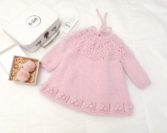 May include: A pink knitted baby dress with a lace pattern and pom-poms around the neckline and hem. The dress has a tie at the back of the neck.