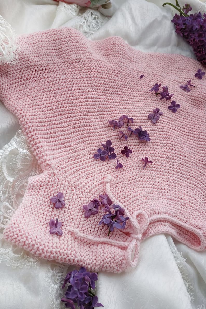 Puede incluir: Una prenda de punto rosa para beb&eacute; con un lazo en el cuello, decorada con flores de lila p&uacute;rpura.