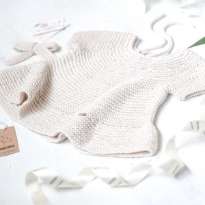 Puede incluir: Un body de beb&eacute; de punto de color crema con un lazo en la espalda. El body est&aacute; hecho de una tela suave y texturizada y tiene un dise&ntilde;o sencillo.