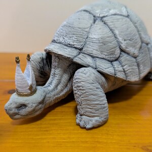 Puede incluir: Una figurita de tortuga de cer&aacute;mica gris que lleva una corona blanca y dorada. La tortuga camina hacia adelante con la cabeza y las patas extendidas.
