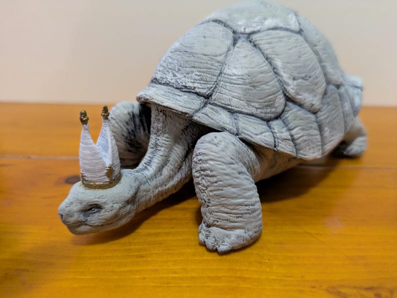 Puede incluir: Una figurita de tortuga de cer&aacute;mica gris que lleva una corona blanca y dorada. La tortuga camina hacia adelante con la cabeza y las patas extendidas.