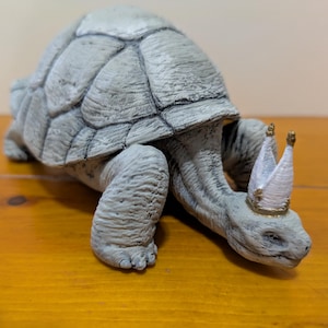 May include: A gray tortoise figurine wearing a white crown with gold accents. The tortoise is walking forward with its head down.