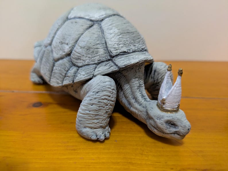 Puede incluir: Una figurita de tortuga gris que lleva una corona blanca con detalles dorados. La tortuga camina hacia adelante con la cabeza baja.