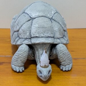 Puede incluir: Una figurita de tortuga de cer&aacute;mica gris que lleva una corona blanca y dorada. La tortuga est&aacute; sentada sobre una superficie de madera.
