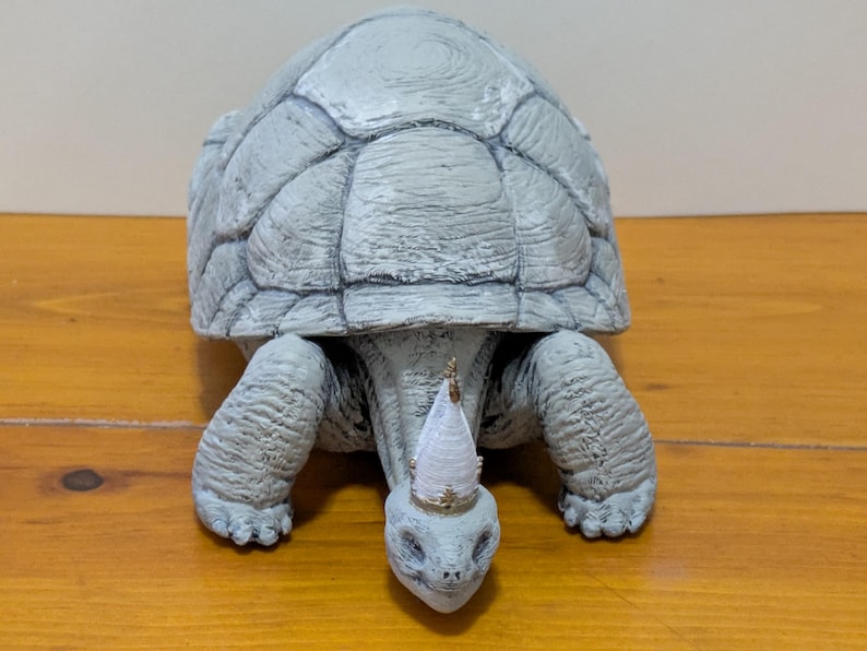 Puede incluir: Una figurita de tortuga de cer&aacute;mica gris que lleva una corona blanca y dorada. La tortuga est&aacute; sentada sobre una superficie de madera.
