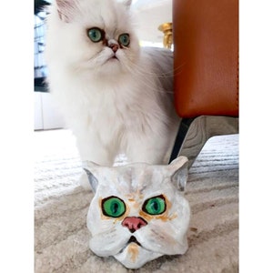 May include: A white Persian cat with green eyes looks at a white ceramic cat head with green eyes. The cat head has a pink nose and a painted expression of surprise.
