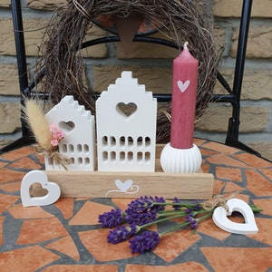 May include: A decorative arrangement featuring white wooden house-shaped candle holders with heart cutouts, a pink candle with a heart, and lavender sprigs. The arrangement is on a mosaic tile table.