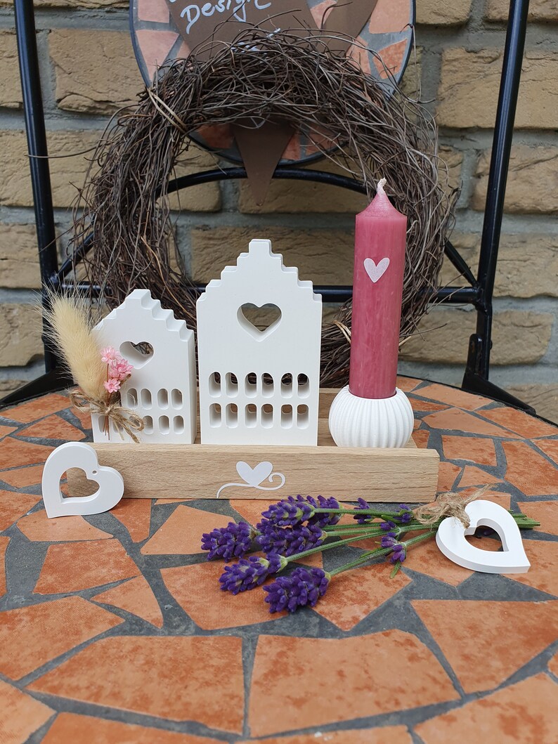 May include: A decorative arrangement featuring white wooden house-shaped candle holders with heart cutouts, a pink candle with a heart, and lavender sprigs. The arrangement is on a mosaic tile table.