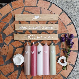 May include: A collection of decorative items, including five pillar candles in shades of beige, red, white, pink, and green, each with a heart design. Wooden signs with the phrases "home sweet home" and "HYGGE zeit" are also present, along with a small white dish and lavender.