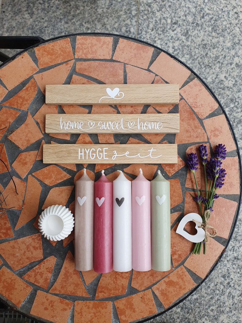 May include: A collection of decorative items, including five pillar candles in shades of beige, red, white, pink, and green, each with a heart design. Wooden signs with the phrases "home sweet home" and "HYGGE zeit" are also present, along with a small white dish and lavender.